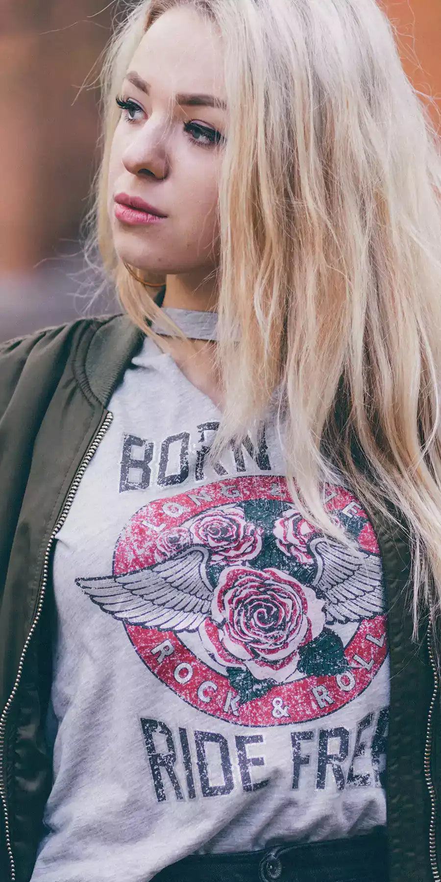 Blond woman wearing an olive colour bomber jacket, underneath a grey t-shirt with a print. The print is roses with wings into a red circle with text - Long live rock & roll. Additional text top and bottom - Born to ride free - mobile