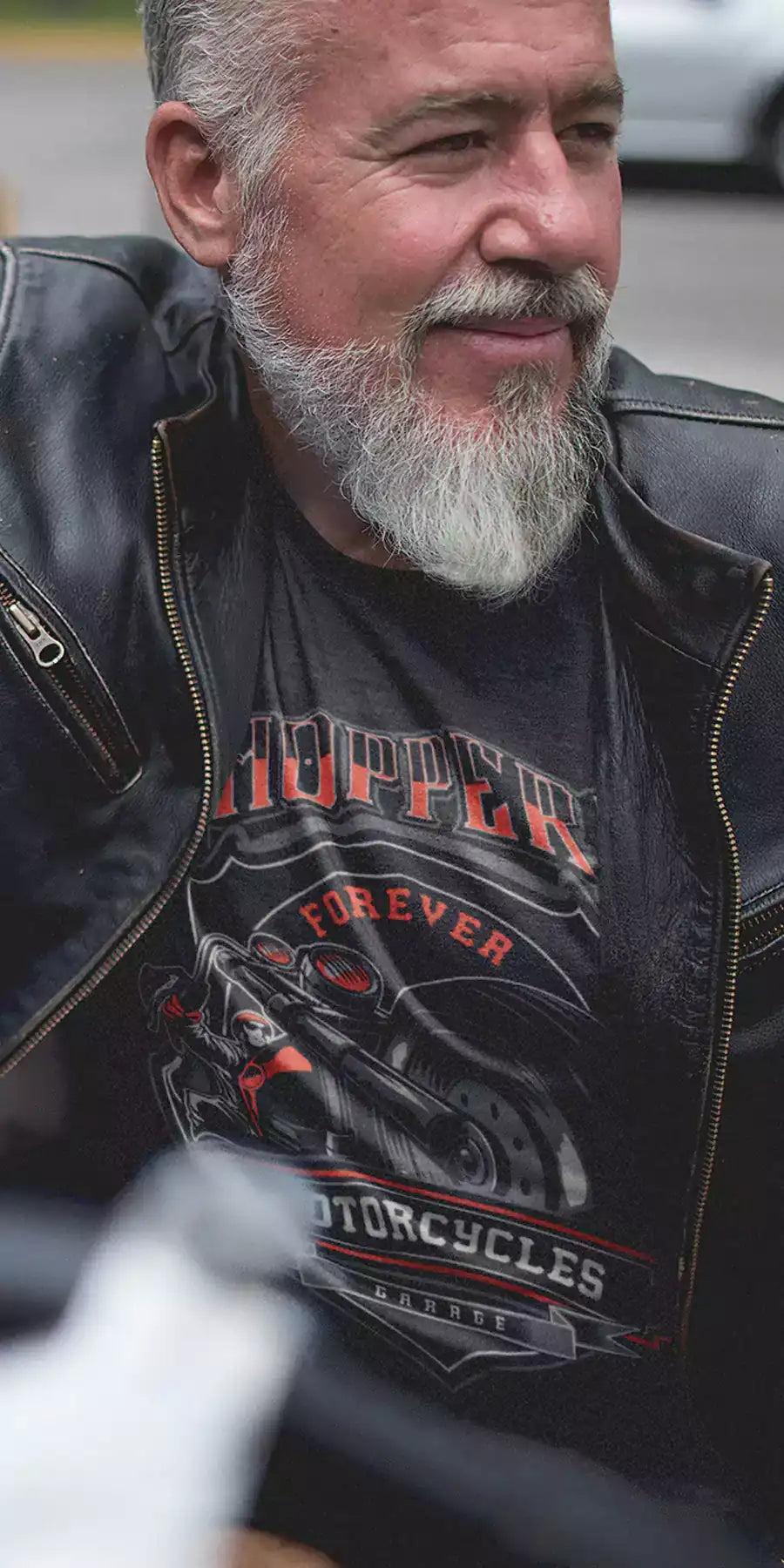 Old man sitting on a motorcycle, wearing a black leather jacket and printed black t-shirt. The t-shirt design is a motorcycle with text - Chopper forever - Mobile