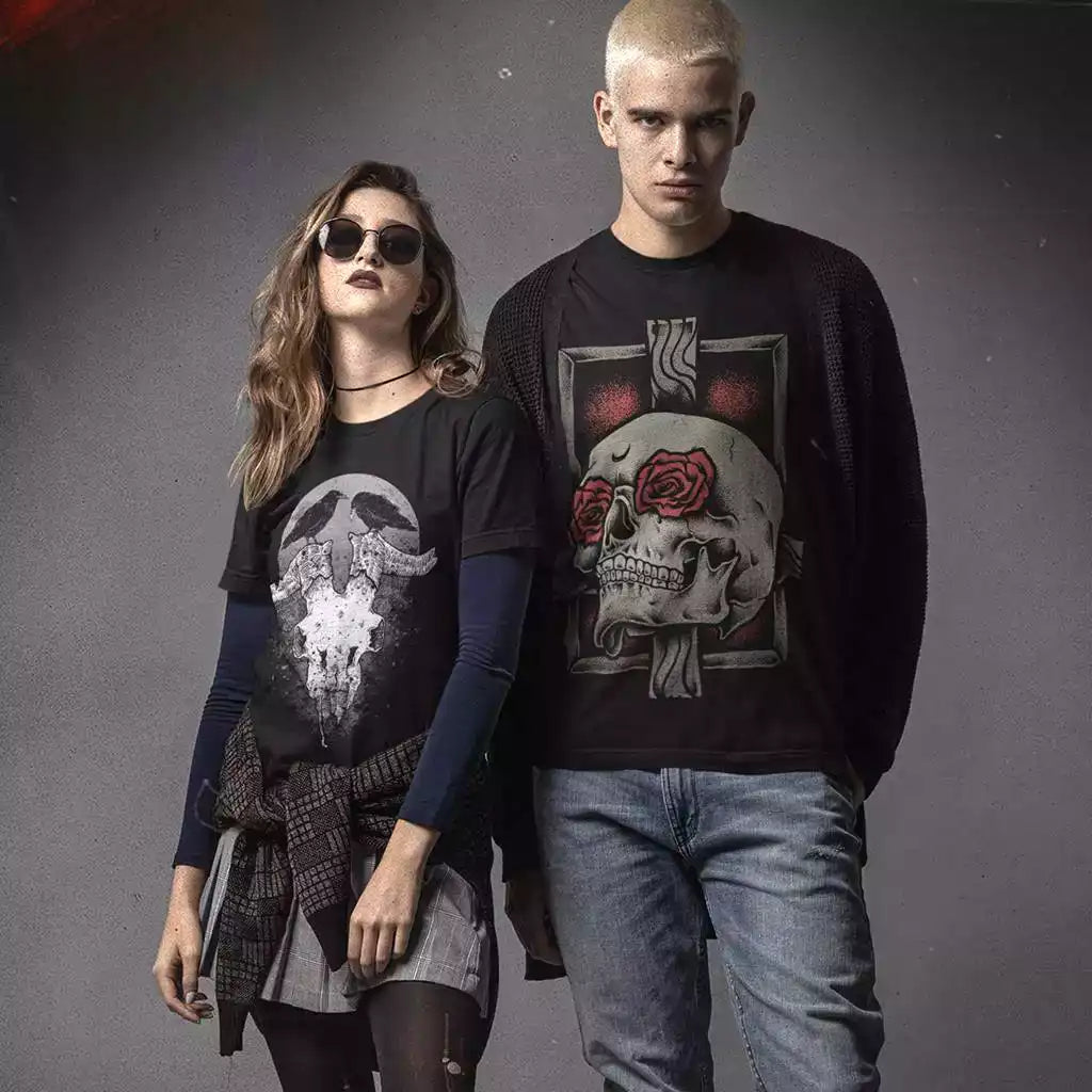 Young man and woman wearing printed black t-shirts. The design on the women's t-shirt is two crows sitting on the horns of an animal skull. The design on the men's t-shirt is a human skull with two roses as eyes and a wooden cross behind