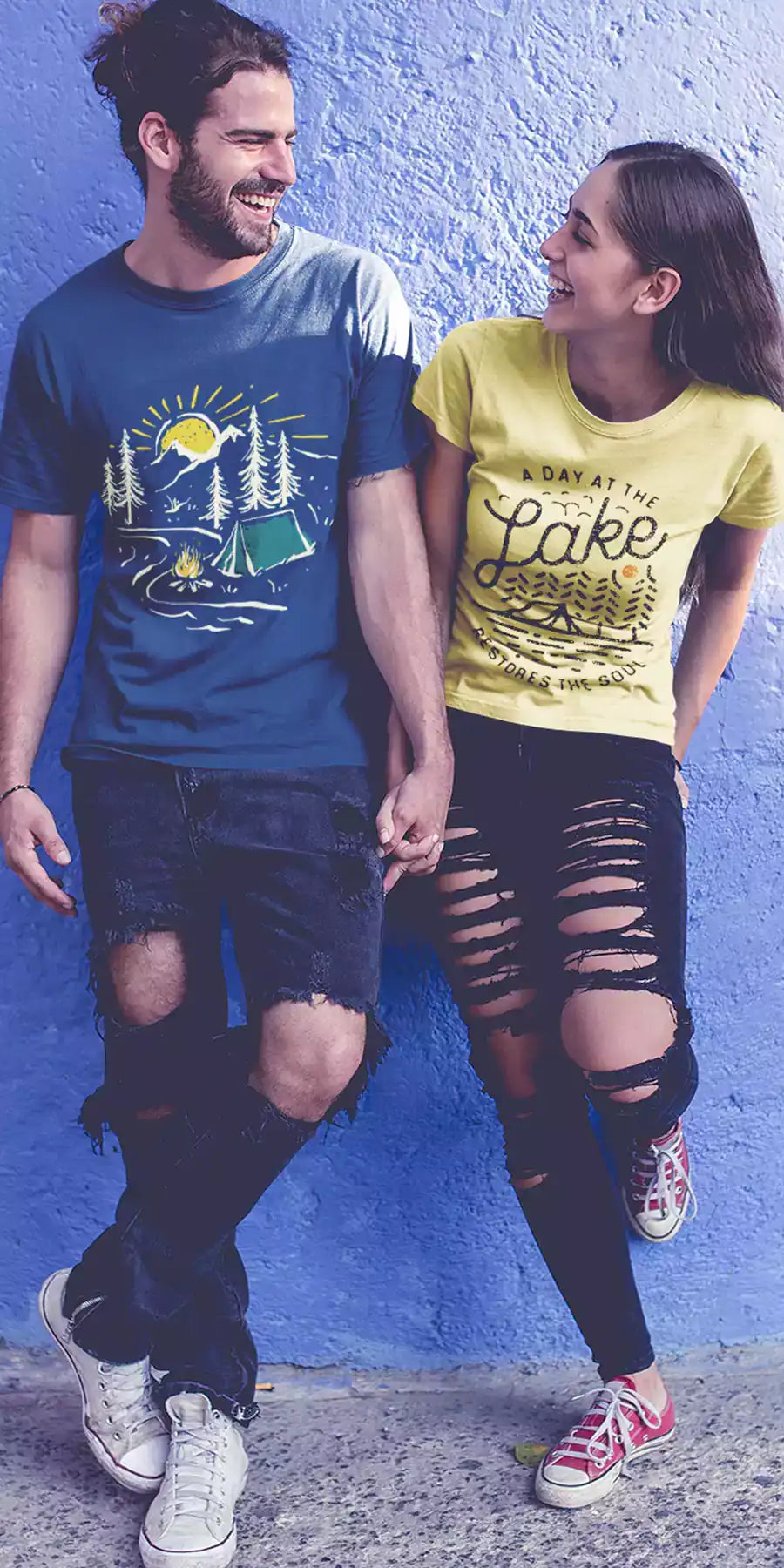 Young man and woman wearing printed t-shirts. The design on the women's yellow t-shirt retro lines graphic and text - A day at the lake. The design on the men's t-shirt is a retro graphic with mountains, sun, river, tent in the forest and a burning fire