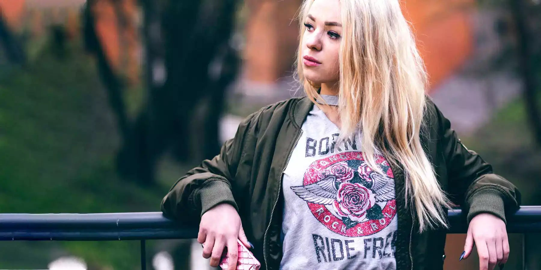 Blond woman wearing an olive colour bomber jacket, underneath a grey t-shirt with a print. The print is roses with wings into a red circle with text - Long live rock & roll. Additional text top and bottom - Born to ride free