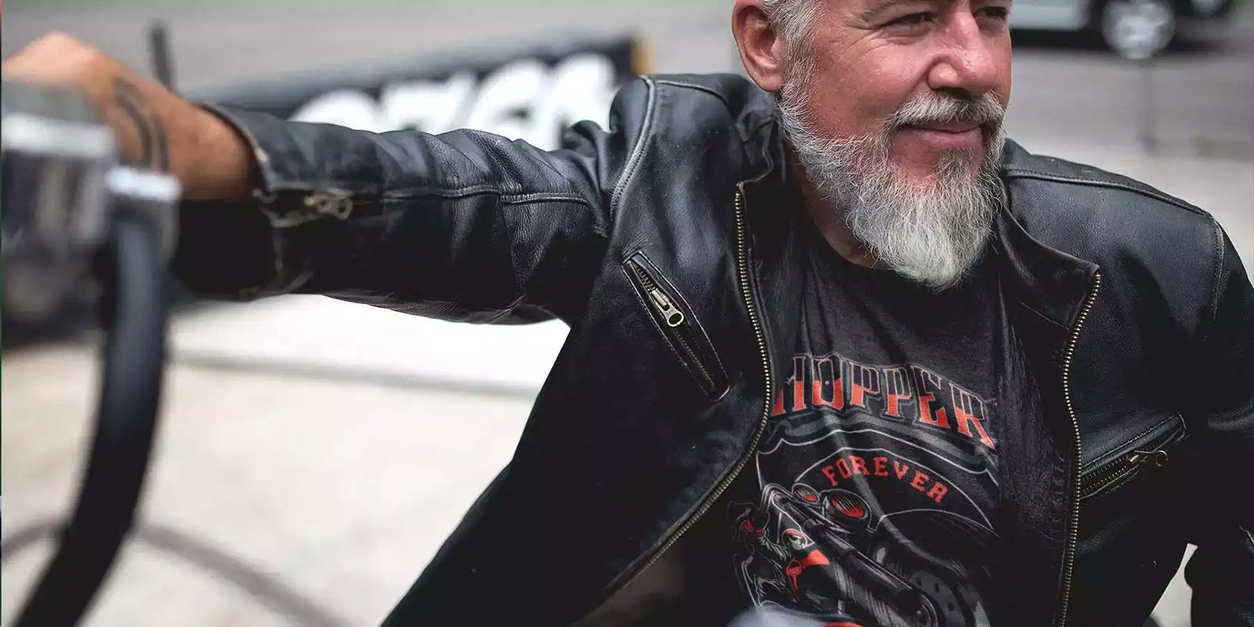 Old man sitting on a motorcycle, wearing a black leather jacket and printed black t-shirt. The t-shirt design is a motorcycle with text - Chopper forever