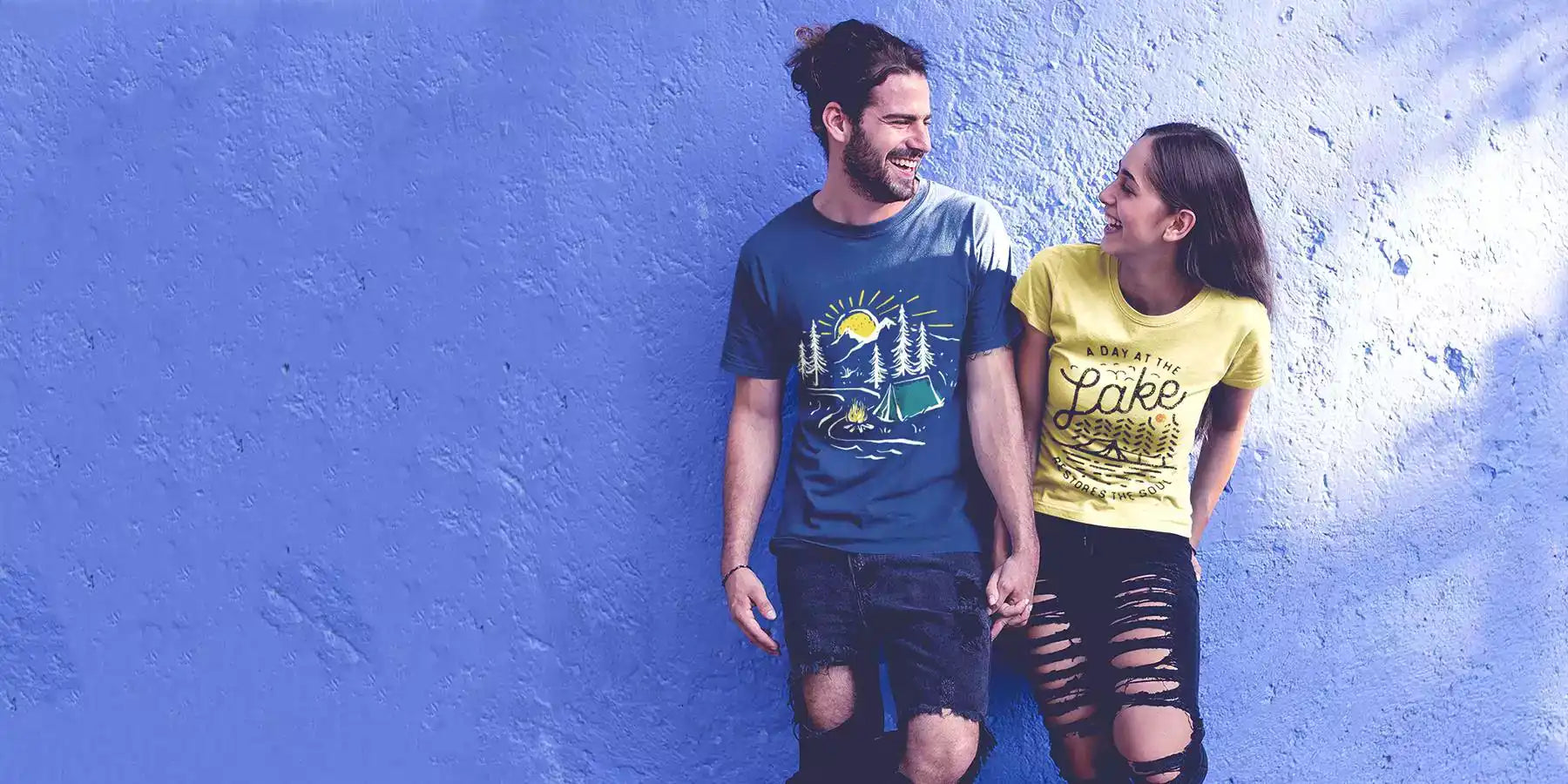 Young man and woman wearing printed t-shirts. The design on the women's yellow t-shirt retro lines graphic and text - A day at the lake. The design on the men's t-shirt is a retro graphic with mountains, sun, river, tent in the forest and a burning fire.