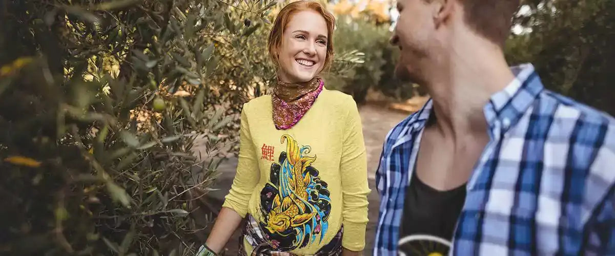 Young man and woman walking between olive trees.  The woman is wearing a yellow long sleeve t-shirt with a Japanese koi fish on the front